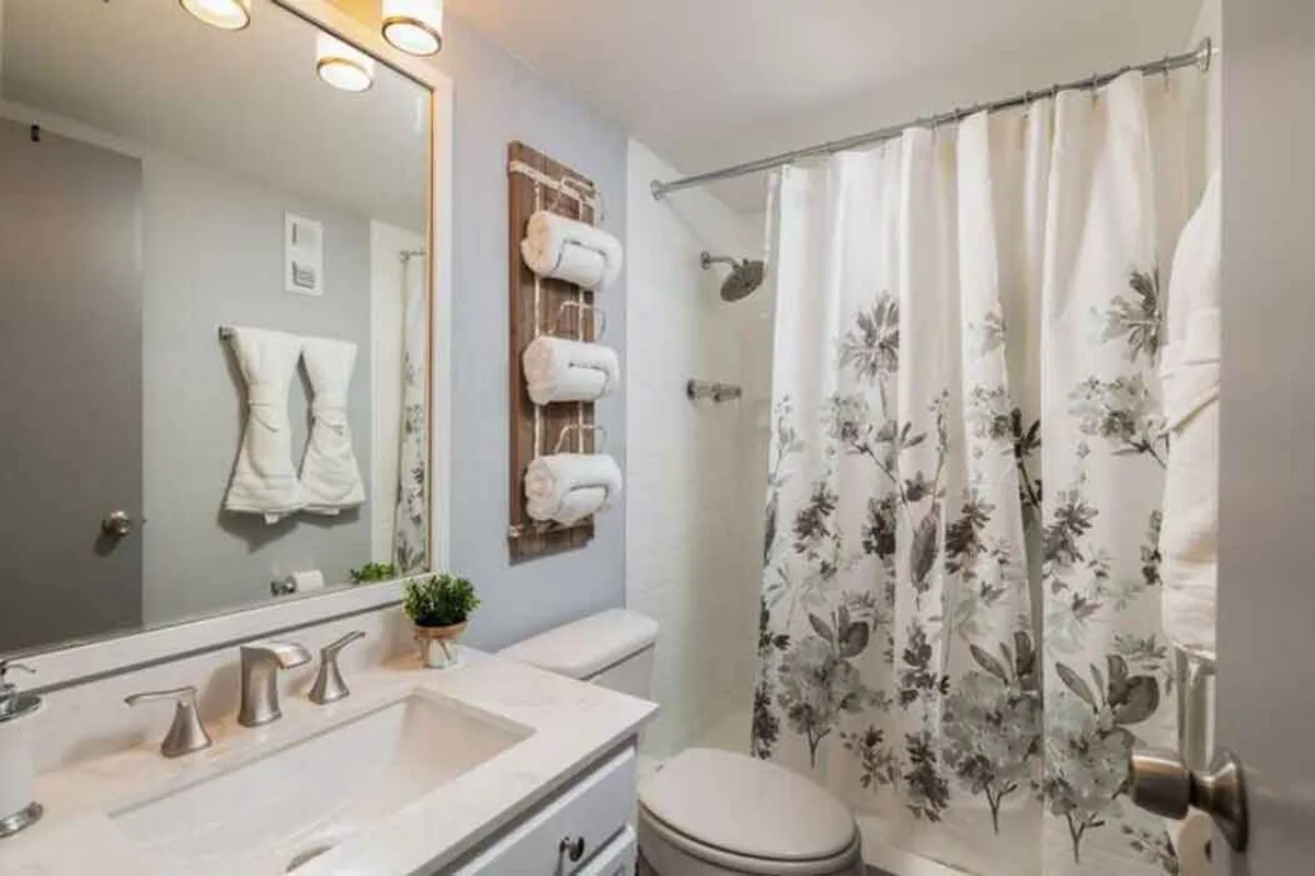 A well-appointed bathroom featuring a vanity with a modern sink, a decorative mirror, a floral shower curtain, towel rack with neatly rolled towels, and a small potted plant. The space feels calm and rejuvenating, ideal for private moments of self-care.