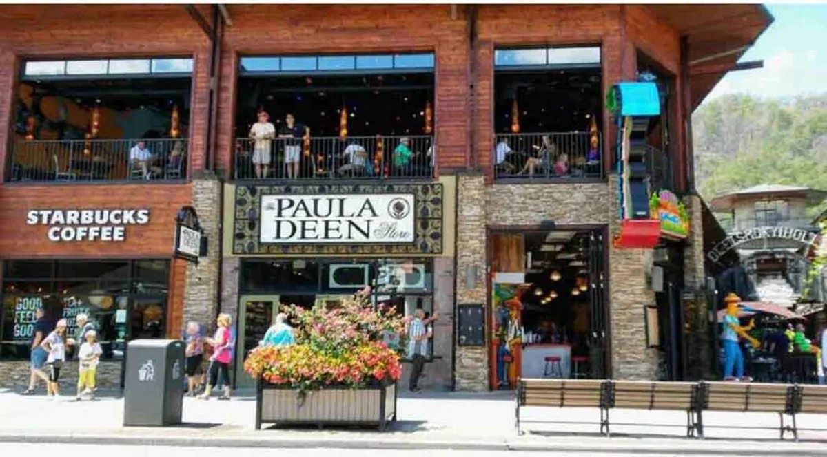 A vibrant streetscape featuring the Paula Deen Store and Starbucks Coffee with flower arrangements, people socializing on a balcony, and an inviting outdoor seating area. The atmosphere is lively, highlighting opportunities for dining, shopping, and enjoying Gatlinburg’s bustling scene.