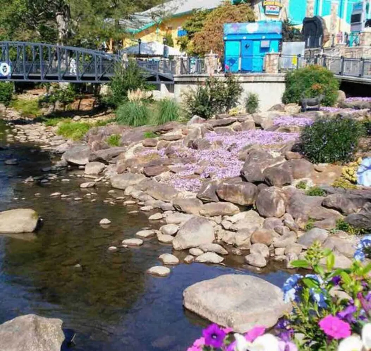 A charming riverside scene with a clear stream winding among rock formations, flower beds in full bloom, a decorative bridge, and cheerful, colorful buildings in the background. People can be seen strolling and exploring nearby shops and nature.