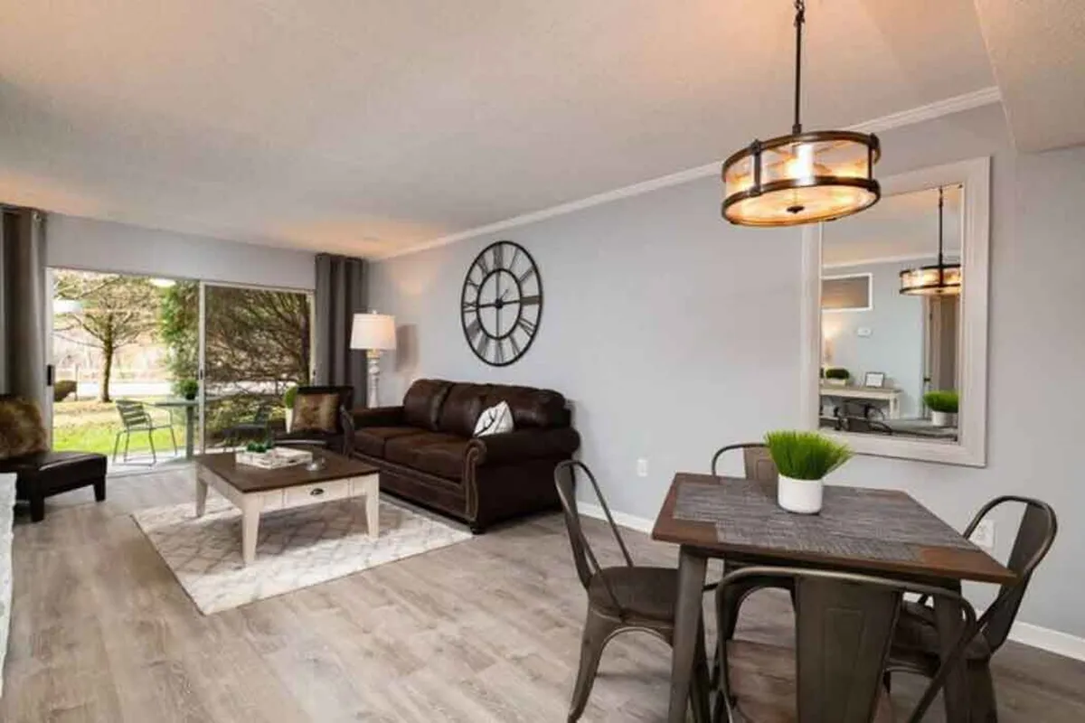 A spacious open-plan living and dining area with a comfortable sofa, a dining table set for meals, a decorative clock, and a large window offering natural light.