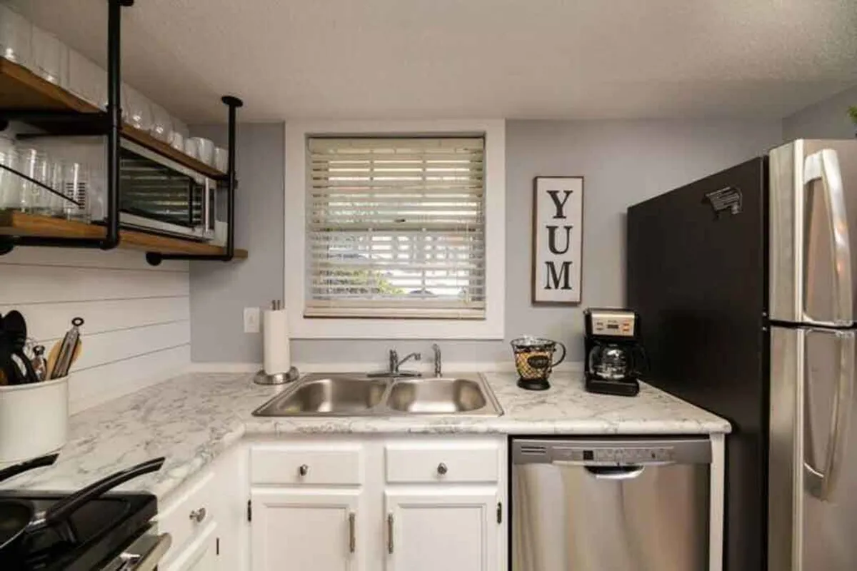 A bright and welcoming kitchen featuring a long marble countertop, stainless steel appliances, open shelving with glassware, and a decorative 'YUM' sign. The space looks modern and fully equipped, with inviting touches that signal both function and comfort.