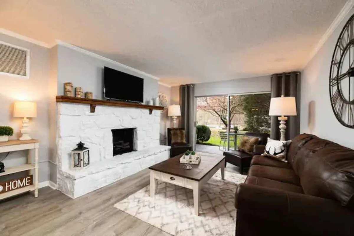 A cozy living room featuring a soft sofa, a crackling fireplace as the centerpiece, a coffee table, large window letting in natural sunlight, and decor that adds warmth to the space.