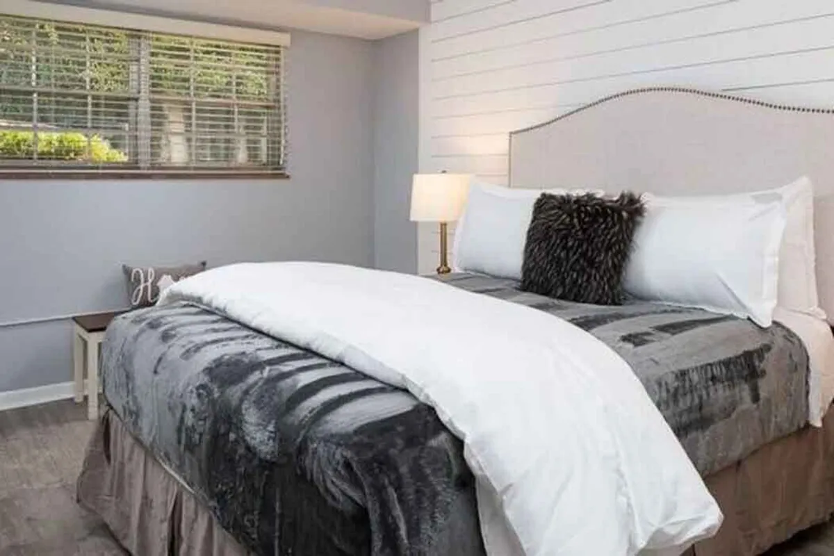 A softly lit bedroom featuring a comfortable bed with plush bedding, a decorative lamp on the nightstand, and a window letting in views of greenery. The room exudes a calm, serene atmosphere perfect for rest and relaxation.