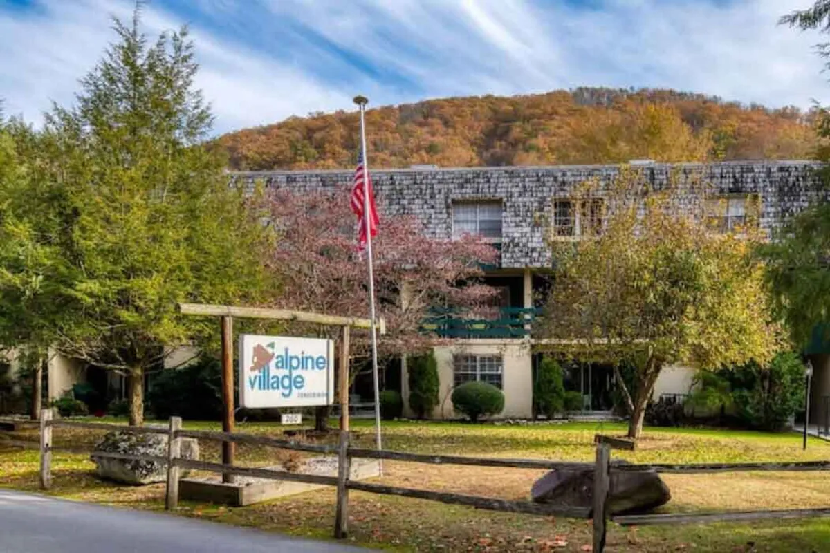 This image shows the front facade of the building, with the 'Alpine Village' sign prominently displayed, a flagpole with the American flag, and attractive landscaping with surrounding trees. The entrance is welcoming, with well-kept greenery giving a sense of serenity and invitation.