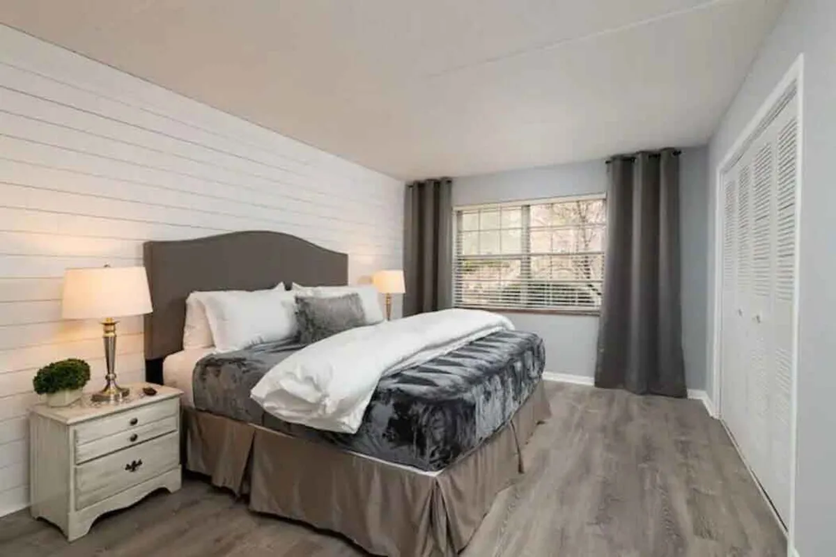 A spacious bedroom with a gray upholstered headboard and matching bedding, a nightstand with a glowing lamp, and a large window bathed in natural light. The room feels comfortable, airy, and inviting.
