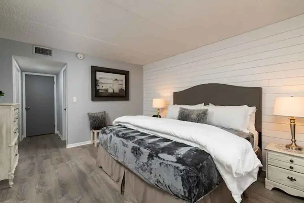 A serene interior bedroom scene featuring a bed with plush bedding, framed artwork, a nightstand with a lamp, and a cozy chair. The mood is calm and restful, suggesting a perfect haven to unwind after busy days.